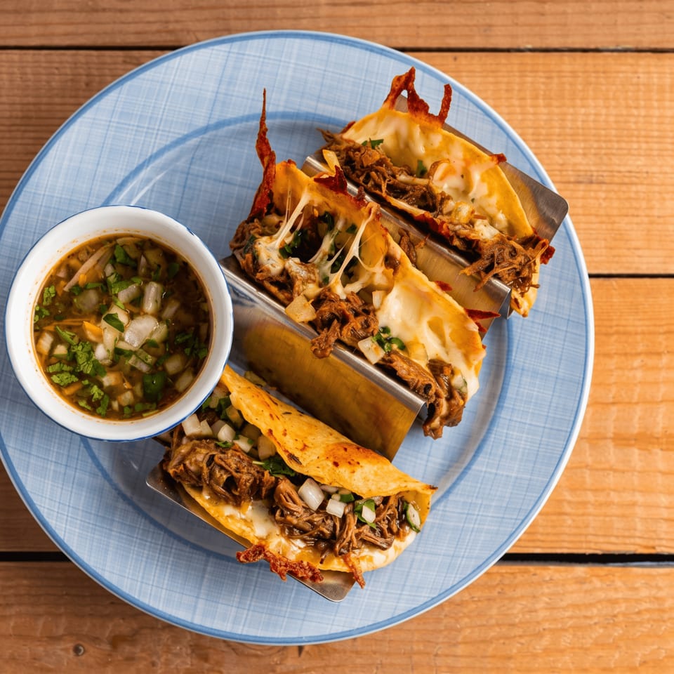 Three cheesy birria tacos with a side of consommé on a blue plate.